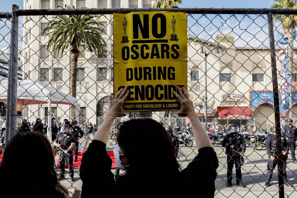 A protester holds a sign during a demonstration in support of Palestinians calling for a ceasefire in Gaza, behind a fence securing the perimeter next to the Dolby Theatre where the 96th Academy Awards Oscars ceremony is held, Sunday, March 10, 2024, in the Hollywood section of Los Angeles. (AP Photo/Etienne Laurent)
